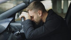 A man showing stress while sitting in a car, head on steering wheel.